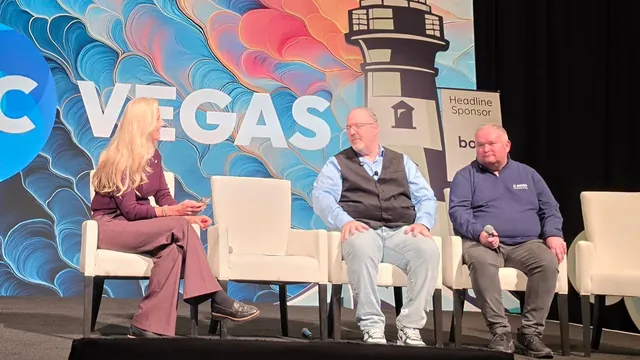 A moderator and two panelists seated on white chairs on the ITC Vegas stage, in front of a colorful lighthouse backdrop.