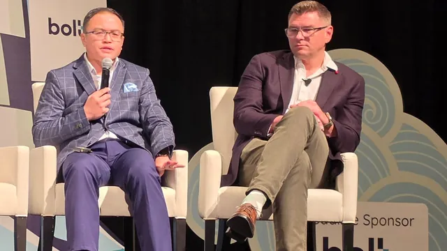 Two panelists seated on stage, one speaking into a handheld microphone while the other listens.