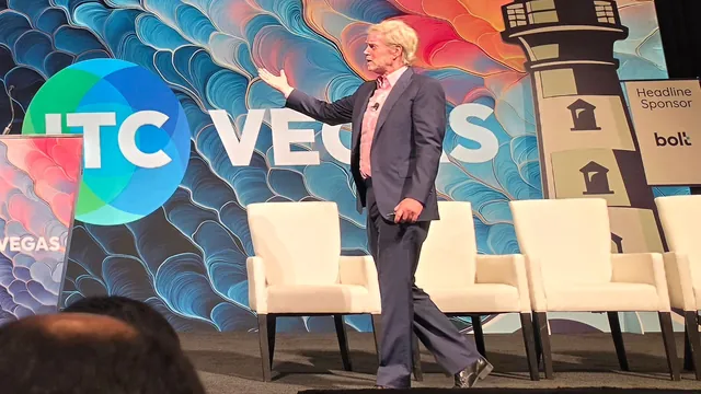 A presenter walking across the ITC Vegas stage, gesturing toward the screen.