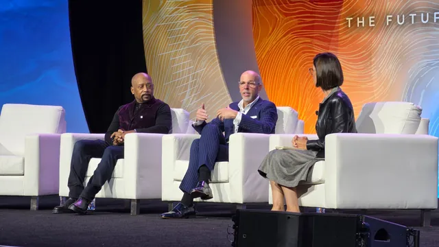 Two guests and a moderator seated in white armchairs on the Main Stage, one guest gesturing while speaking.